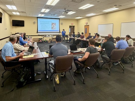 A large room featuring individuals gathered around a square of multiple tables attending a training event.