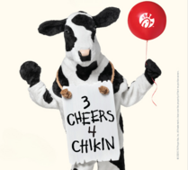 Chick-fil-A cow mascot holds a red Chick-fil-A balloon and wears a sign reading, “3 Cheers 4 Chikin.”
