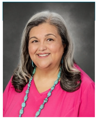 a smiling woman with long, wavy salt-and-pepper hair. She is wearing a bright pink top, turquoise beaded jewelry