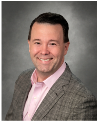 a smiling man with short dark hair. He is wearing a pink collared shirt and a gray checked blazer