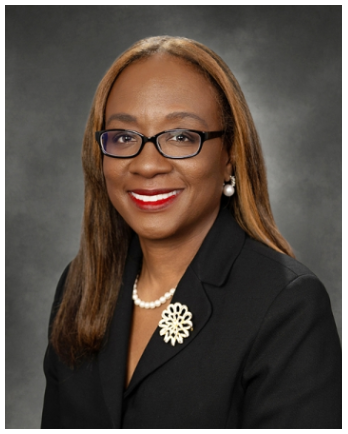 a smiling woman with long, straight brown hair and glasses. She is wearing a black blazer