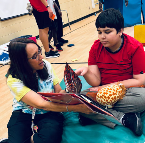 The image shows an adult woman sitting beside a student looking at a book