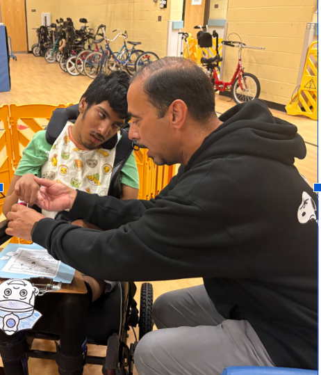 The image shows an adult man in a black hoodie assisting a seated young person in a wheelchair