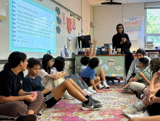 Fifth-grade students sit in a circle on the classroom rug and participate in a morning meeting with their teacher.