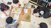 students working on the floor with paper and pencil with a bucket that has corn in it placed in the middle of the students