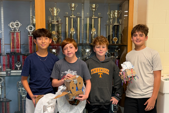 Four students pose with their egg-drop designs in front of a school trophy case.