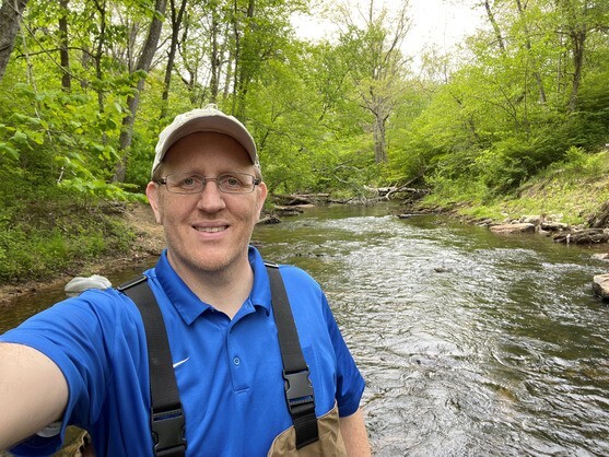 Bradley Webster wearing fishing waders and standing outdoors in front of a forest stream.