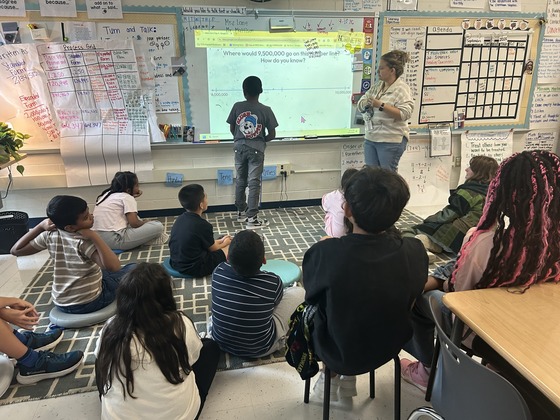 A student solves a math problem on the whiteboard while his teacher and classmates look on.
