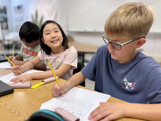 Three third-grade students write with pencils at a classroom table.