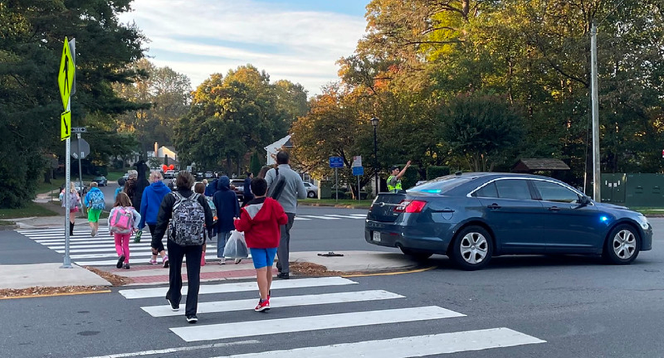 a photo of adults and students crossing the street at a crosswalk with a police officer holding traffic for them.