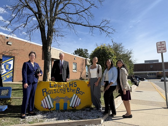 Three adults and two students standing beside yellow stone with the motto Raising the Bar in front of a school building.