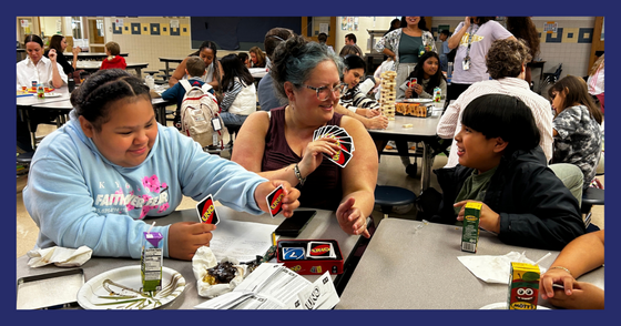 Two students and a teacher play UNO at a table in the cafeteria. Everyone is smiling and laughing.
