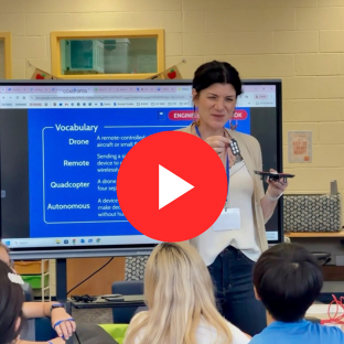 Woman standing in front of TV with drone vocabulary on it holding a drone and battery to show to students. Red play button in the center of the image