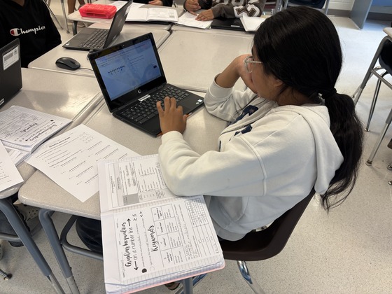A student sits at her desk with her computer and a notebook