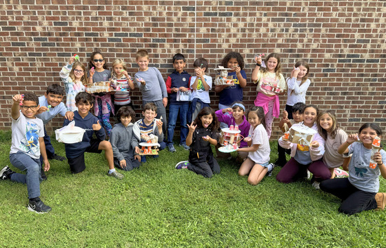 A group of elementary school children poses outdoors on the grass in front of a brick wall, holding up handmade craft structures.