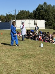 students sitting on grass. man in science coat and woman in blue suit standing in front of them