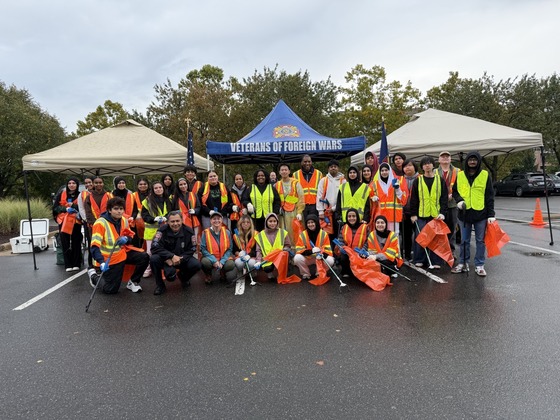 Students and teachers in orange and yellow vests in front of a tent that says Veterans of Foreign Wars with cleaning tools and orange trash bags