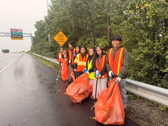 Students on highway in yellow and orange vests with cleaning tools and orange trash bags