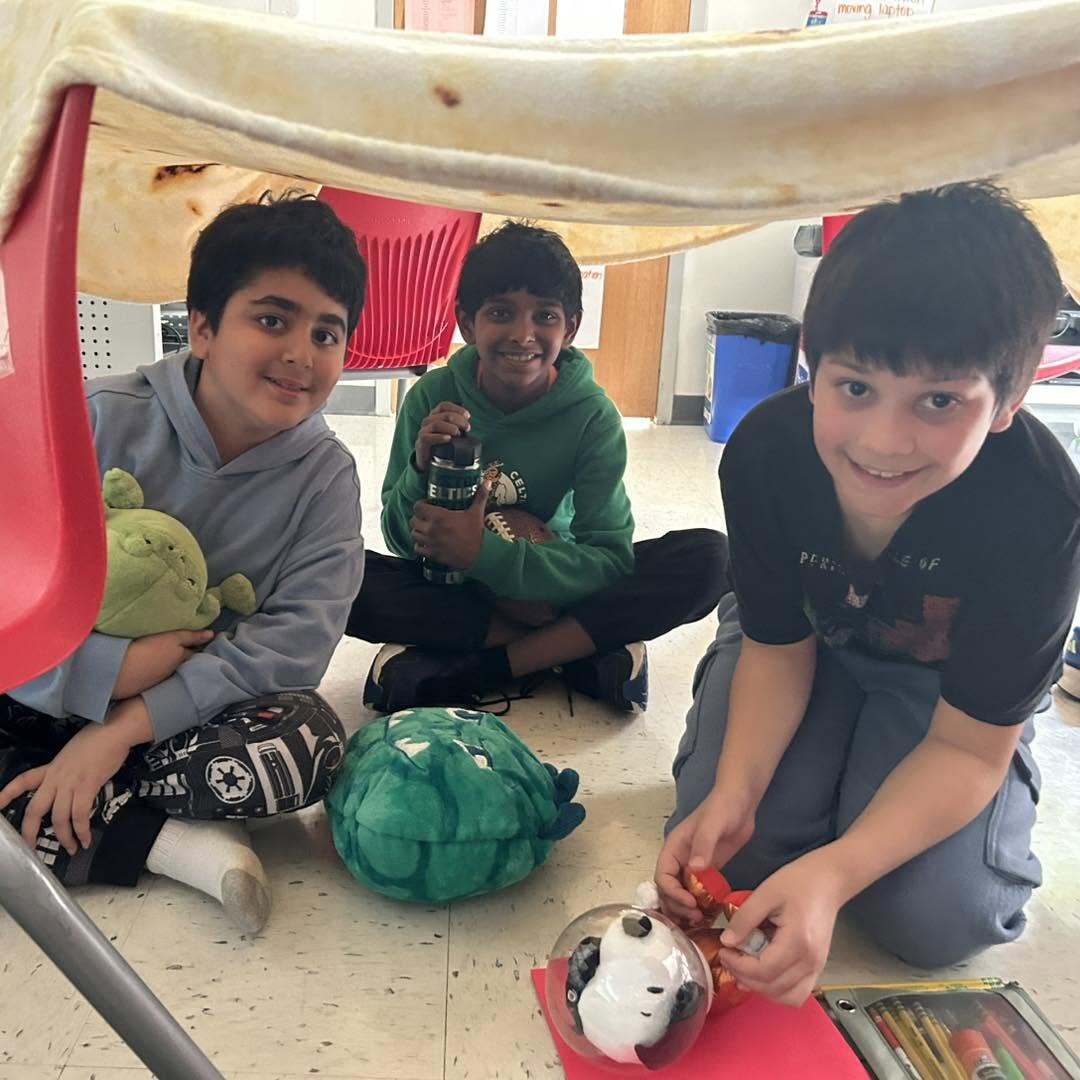 Three fifth grade students smile at the camera while sitting under their desks in a fort