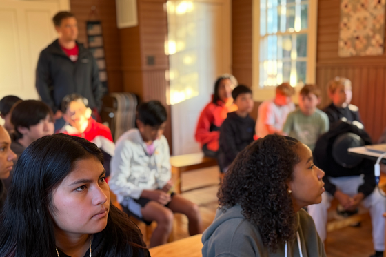 Students sit on wooden benches inside the Crouch School House as an adult speaks to the group.