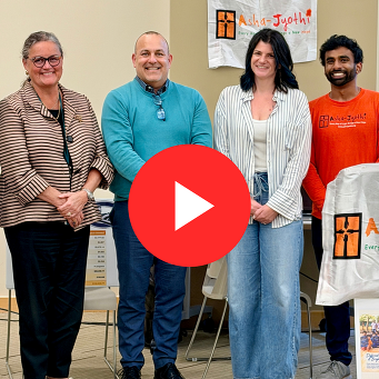 Image of 4 adults smiling for a photo with the Asha Jyothi logo hanging in the background. There's a red play button over the center of the image
