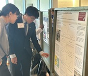 A student dressed in a suit shows off a research poster to a parent