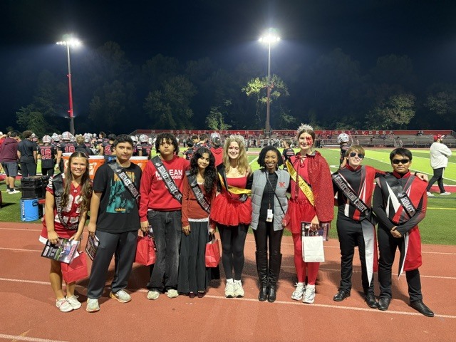 McLean High School homecoming court smiles on the stadium track under bright night lights wearing red attire and sashes.