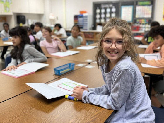 A student smiles at her desk with an open math notebook during a classroom lesson.