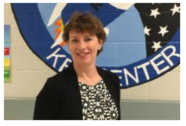 A smiling woman with short brown hair stands in front of a large blue-and-white school emblem, rockets with stars.png