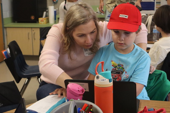 A 2nd grader wearing a blue shirt and a red cap on the computer showing his parent his work