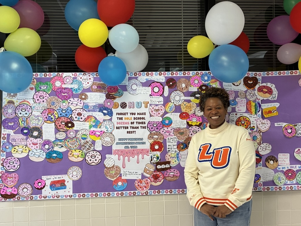 Ms. Yarn smiling in front of a colorful donut-themed bulletin board for Principal Appreciation Month.