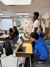 Sudents in a classroom participating in a debate with open laptops on their desks, while a timer counts down on a projection screen in the background.
