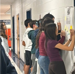 English 8 students standing in a hallway gallery walk, writing notes on sticky pads posted on large poster sheets along the wall.