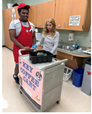 Student wearing a red apron and cap pushing a mobile cart labeled "Key Coffee Cart" alongside a staff member helping prepare drink orders.