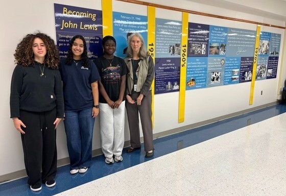 Three library student interns posing alongside volunteer in front of a wall timeline display titled "Becoming John Lewis."