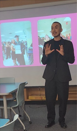 Alumnus Axel Vasquez gesturing with his hands while speaking to students in front of a presentation screen featuring his graduation photos