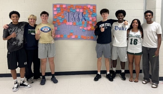 Student leaders standing together in front of a poster displaying "988 National Suicide Hotline" surrounded by colorful post-it notes of support.