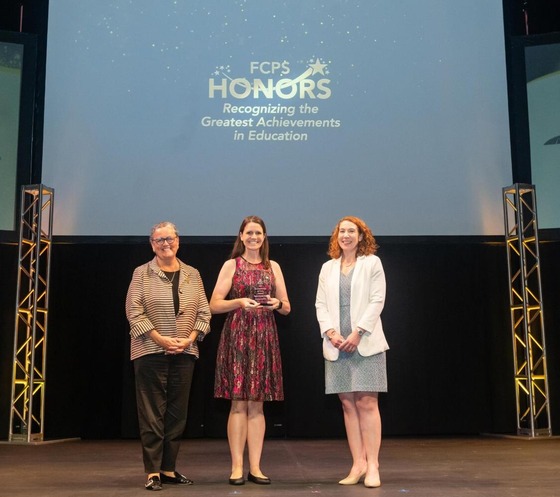 White Oaks Elementary teacher Megan Haile standing on stage holding an award, flanked by two women