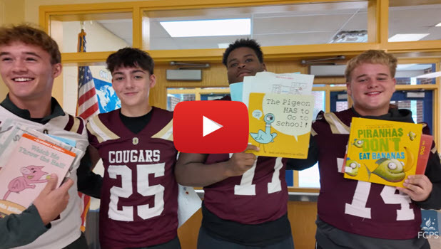 High school football players in Cougar football jerseys stand side-by-side inside a school hallway while holding children's picture books