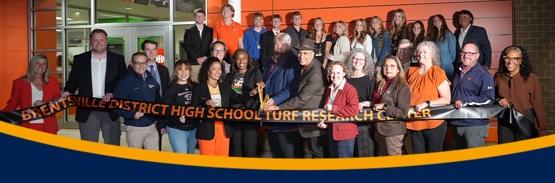 leaders and students holding long "Brentsville District High School Turf Research Center" ribbon and cutting with golden scissors