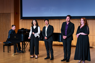 Man seated playing the piano with four male and female singers standing to his right and facing the audience 