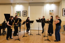 6 women stand, dressed in black, and playing saxophones, flutes and a clarinet 