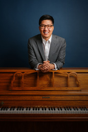 Man smiling and standing behind an upright piano made of brown wood. 