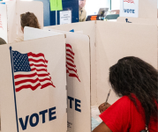 Person voting behind a voting divider that has the text "Vote" and an image of an American flag.