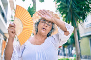 Photo of an older woman outdoors in hot weather, holding a fan and wiping her forehead.