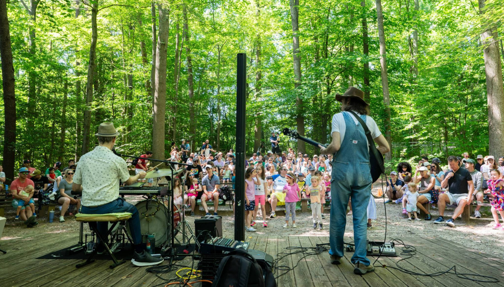 Image of a two-man band performing on a wooden stage in the middle of a green forest, performing to an audience of kids and parents.