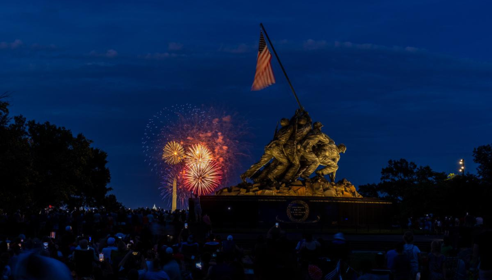 The Marine Corps War Memorial with an American flag is illuminated at night. Fireworks burst in the sky near the Washington Monument. 
