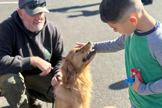 Young boy pets golden retriever type dog as it happily lifts its head and opens mouth. Man squats down behind dog, holding its leash and smiles.