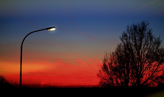 A glowing streetlamp stands against a vibrant sunset sky transitioning from red and orange hues to deep blue. Silhouetted trees frame the scene.