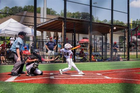 A child swings dress in a baseball uniform and helmet swings a bat at home plate. 